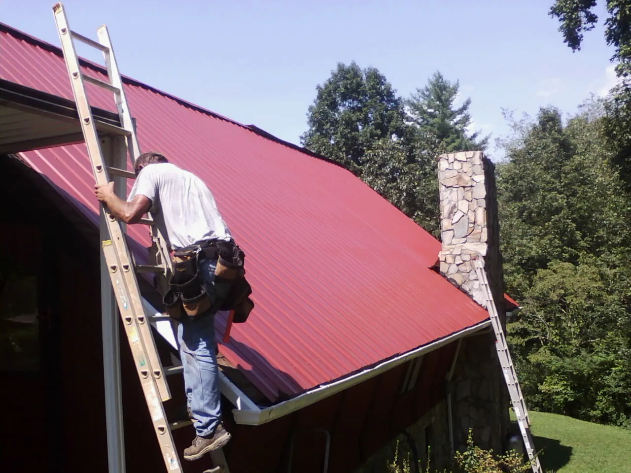 Roofer on a ladder beside a red metal roof and stone chimney — Soffit & Fascia Repair by Roofing Brothers in Canyon Lake